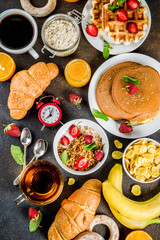 Healthy breakfast eating concept, various morning food - pancakes, waffles, croissant oatmeal sandwich and granola with yogurt, fruit, berries, coffee, tea, orange juice, dark rusty background