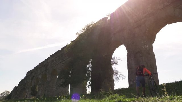 Cyclist with bike arrives under majestic ancient roman aqueduct at sunset. Young attractive athletic man with orange sportswear and backpack in parco degli acquedotti in Rome