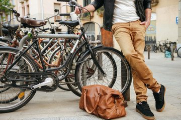 Cropped photo of stylish adult man in casual wear with leather bag standing on city street near bicycle parking