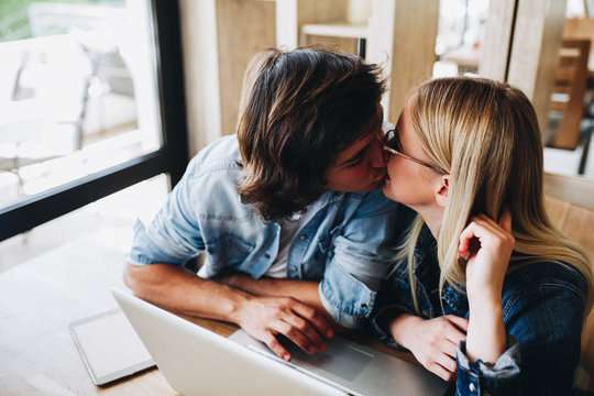 Romantic young couple kissing while sitting at cafe and using laptop