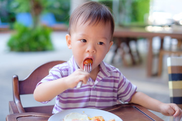 Cute little Asian 18 months / 1 year old toddler boy child sitting in high chair using fork eating...