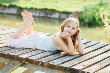 Portrait of cute blond 7 years old girl lying on the wooden bridge at the summer time.