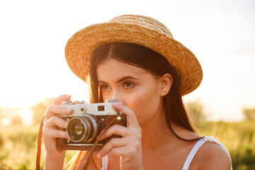 Closeup picture of beautiful young woman with long dark hair wearing straw hat photographing on retro camera, while resting in sunlit nature