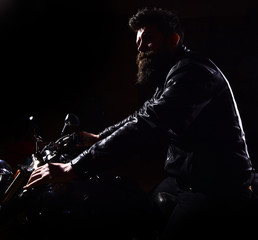 Fototapeta na wymiar Hipster, brutal biker in leather jacket riding motorcycle at night time, copy space. Night rider concept. Man with beard, biker in leather jacket sitting on motor bike in darkness, black background.