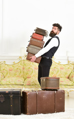 Man with beard and mustache wearing classic suit delivers luggage, luxury white interior background. Butler and service concept. Macho, elegant porter on strict face carries pile of vintage suitcases.