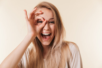 Image of amusing caucasian woman wearing casual clothing laughing and looking at you through hole made by fingers like ok sign, isolated over beige background