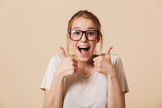 Photo of excited woman 20s with blonde tied hair wearing basic t-shirt and eyeglasses laughing while showing thumbs up, isolated over beige background in studio