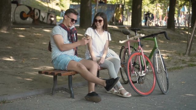Smiling couple discussing taken selfie near bikes while sitting on the bench in lovely green park, relaxing outdoors on hot summer day