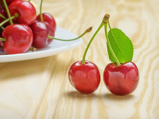 Two ripe red cherries with a green leaf next to a white plate filled with cherries. Light wood background