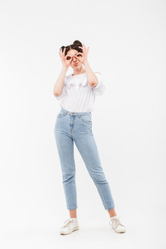 Full length portrait of funny cheerful girl 20s with two buns hairstyle looking at you through binoculars making with fingers, isolated over white background in studio
