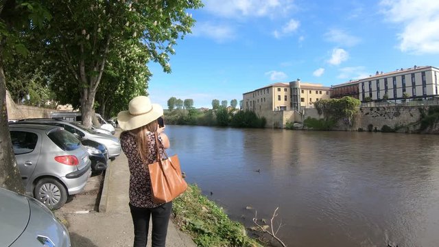 Young woman with a hat and a handbag photographing with her smartphone and strolling along the river Aude in the French town of Limoux, Languedoc in the south of France