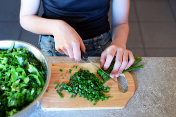Teenage girl hands cutting fresh spring onion  with kitchen knife and wooden cutting board for preparing green salad for dinner on grunge grey table. Natural homemade cooking concept.
