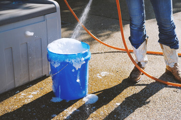 Summer lifestyle scene with woman in cowboy boots filling blue bucket of soapy water to do cleaning...