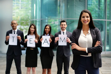 Obraz premium Portrait of a business woman, in a suit and tie, happy and smiling as she looks at the camera and in the background a group of multi-ethnic business people. Concept of: success and female career