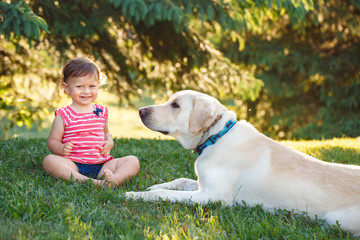 Portrait of cute adorable little Caucasian European baby girl sitting with dog in park outside. Child feeding animal domestic pet. Happy childhood concept