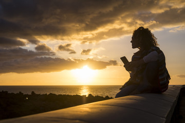 beautiful woman in silhouette during a golden amazing sunset on the ocean using a technolofy mobile smart phone sitting down on a long stone beanch. nice leisure in vacation writing friends