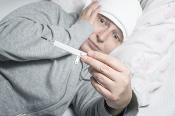Woman with fever checking her temperature with a thermometer at home
