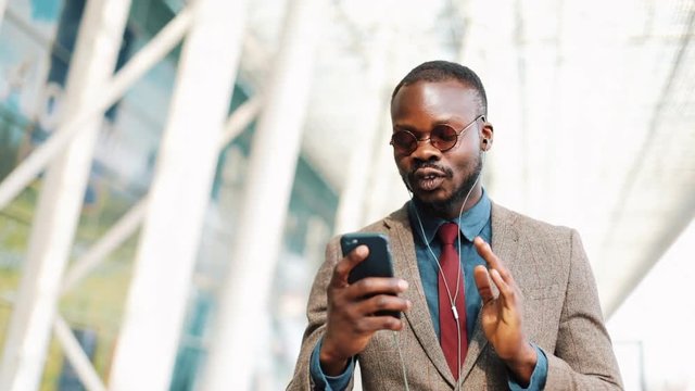 African American businessman in the sunglasses talking on the phone with headphones near the office building. Business, people, music, technology, leisure and lifestyle concept