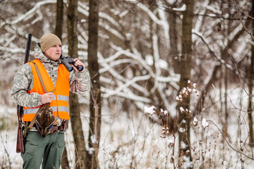 Male hunter in camouflage, armed with a rifle, standing in a snowy winter forest with duck prey