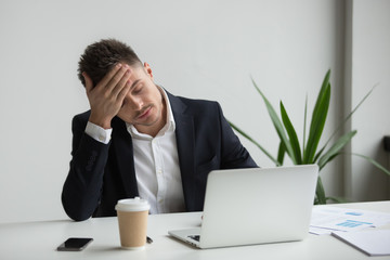 Frustrated fatigued millennial businessman having strong headache tired from laptop use touching...