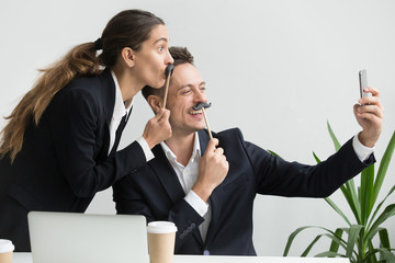 Friendly colleagues in suits having fun grimacing holding fake mustache taking picture on...