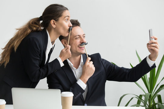 Friendly colleagues in suits having fun grimacing holding fake mustache taking picture on smartphone in office, happy coworkers making funny faces laughing at phone camera, silly selfie concept