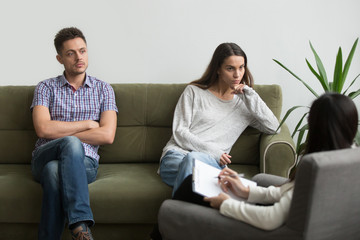 Unhappy millennial couple sitting apart on couch visiting female psychologist listening to counselor helping solving problems in bad relationships, family therapy session, marriage counseling concept