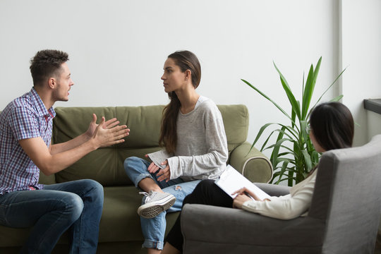 Unhappy couple arguing having fight disagreement at psychologists office, frustrated young family discussing relationship problems sitting on couch during therapy session, marriage counseling concept