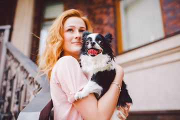 A beautiful young woman with red long hair is holding a small, cute funny big-eyed dog of two flowers, a black-and-white pet of the breed of hichuahua against a house of red brick in summer