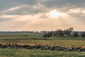 Sunset image of sun peaking through clouds and shining down on red barn in a field on a farm. surrounding by rock fence.