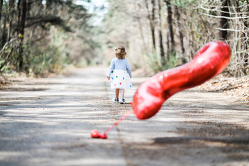 Blonde white female toddler wearing a grey and white dress with multi colored butterflies walking away from a red number two balloon.