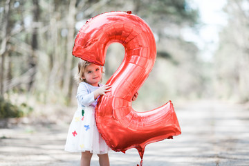 Blonde white female toddler wearing a grey and white dress with multi colored butterflies holding and looking through a red number two balloon smiling at camera.
