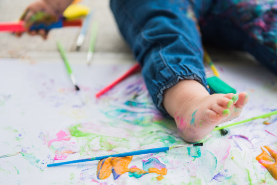 White toddler foot with splattered paint surrounded by paint brushes and paint covered paper.