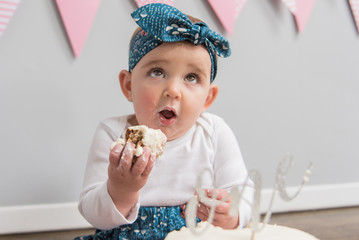 Blonde white female toddler wearing a blue patterned headband and matching shorts with white shirt eating and enjoying cake.