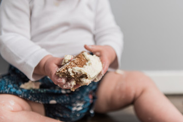 White toddler wearing a blue patterned shorts with white shirt playing with cake in hands.