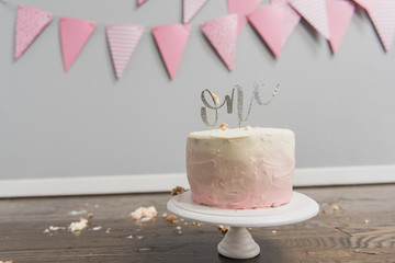 Cream and pink ombre cake on white cake stand with one sign on top with cake pieces on floor and pink and white patterned flag banner in background.