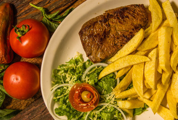 Close up of detailed grilled beef steak with french fried potato, tomato, served in white plate on cutting board on wooden table background