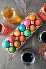 Top view of multi-colored eggs drying in a cardboard egg container on top of parchment paper surrounded by dyed water in mason jars..