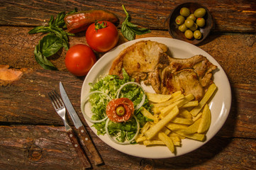 Above view of medium grilled Lamp chop with french potatoes served on white plate, tomatoes,carrots, fork and knife on cutting board, on wooden background