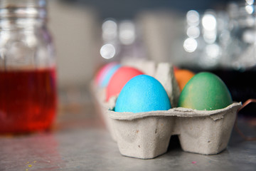 Side view of dyed colored eggs in cardboard egg container with mason jars filled with dyed water in the background.