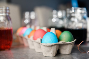 Side view of dyed colored eggs in cardboard egg container with mason jars filled with dyed water in the background.