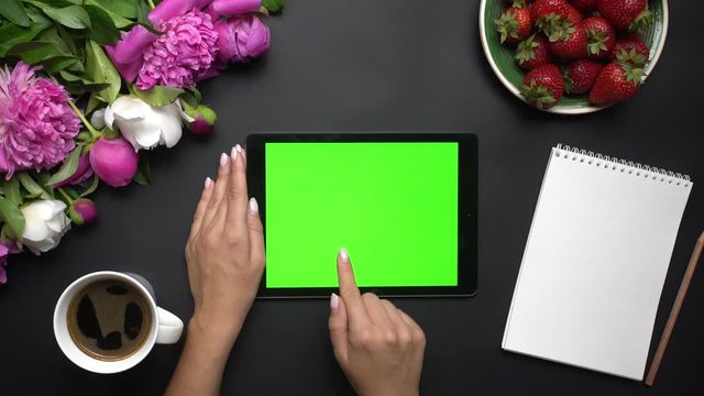 Woman working on tablet computer device with green screen. Beautiful white and pink peonies flowers, strawberries, coffe and notebook on the black background. Table top view. Chroma key