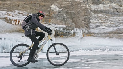 Obraz premium Man is riding bicycle near the ice grotto. The rock with ice caves and icicles is very beautiful. The cyclist is dressed in gray down jacket, cycling backpack and helmet. The tires on covered with