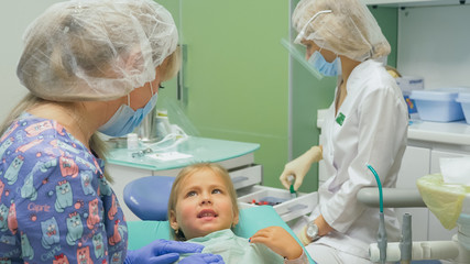 Child with a mother at a dentist's reception. The girl lies in the chair, behind her mother. The doctor works with an assistant. Procedure for drilling a tooth. Setting a temporary seal. Using the x