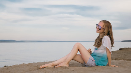 Fototapeta na wymiar One beautiful teenage girl with brown hair outside on a beautiful summer day. The woman is sitting on the beach. The sun was gone in the sunset. The girl is genuinely genuinely genuinely smiling and