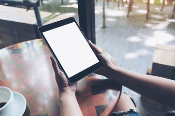 Mockup image of hands holding black tablet pc with white blank screen and coffee cup on table background