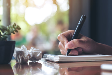 Closeup image of a hand working and writing down on a white blank notebook with screwed up papers on table