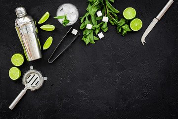 Ingredients and crockery for making mojito. Slices of lime, mint, sugar cubes, glass with ice cubes, shaker, strainer on black background top view copy space