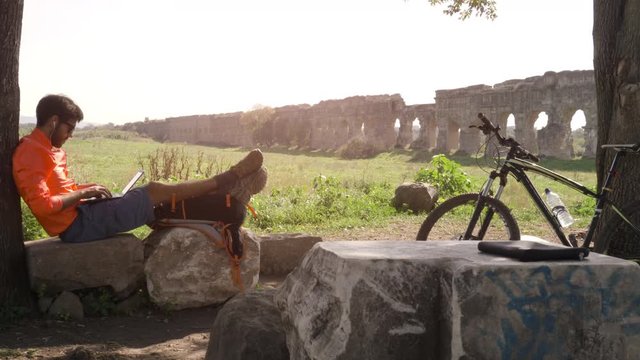 Cyclist with bike sitting on a rock under a tree writing on laptop in front of ancient roman aqueduct. Young attractive athletic man with orange sportswear and backpack in parco degli acquedotti in