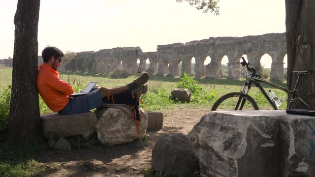 Cyclist with bike sitting on a rock under a tree on a videocall with his laptop in front of ancient roman aqueduct. Young attractive athletic man with orange sportswear and backpack in parco degli